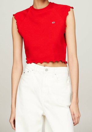 Frau trägt ein rotes, gestuftes, ärmelloses Crop-Top und hochtaillierte weiße Hose, steht vor einem schlichten Hintergrund.