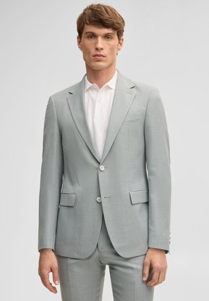 Junger Mann in hellgrauem Anzug und weißem Hemd mit Kragen, vor einfarbigem hellgrauem Hintergrund stehend.