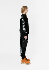 Sudadera negra con texto gráfico blanco en las mangas, combinada con pantalones cargo negros con bolsillos. Accesorios: botas marrones con suelas gruesas.