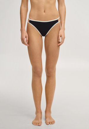 Vrouw die een zwarte bikini-onderbroek met witte rand draagt, blootsvoets staand tegen een effen lichte achtergrond.