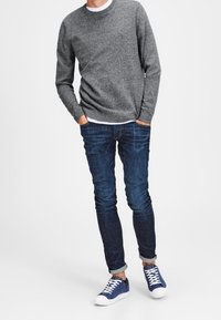 Grå stickad tröja, blå slim-fit jeans och blå sneakers med vita sulor. Tröjan har en rund hals; jeansen är något uppvikta vid fållarna.