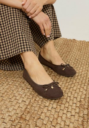 Donna che indossa ballerine in suede marrone con piccoli dettagli a fiocco, seduta su un tappeto in fibra naturale intrecciata, vestita con gonna a quadri marrone.