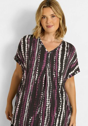 Mujer con cabello rubio hasta los hombros que lleva una camiseta de manga corta en V con rayas verticales en negro, morado y blanco.