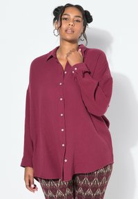 Camicetta button-up borgogna con finitura testurizzata, caratterizzata da maniche lunghe e una vestibilità rilassata. Accentuata da bottoni bianchi e sottili pieghe.