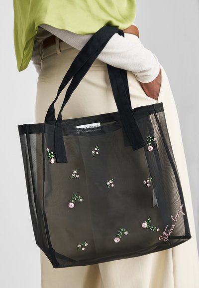 Person, der holder en sort gennemsigtig tote bag med små pink blomsterbroderier og Stella McCartney-logo nær bunden.