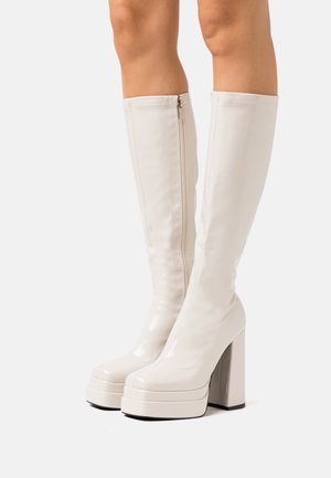 Bottes blanches montantes jusqu'aux genoux avec plateformes, talons épais et zips latéraux, portées sur des jambes nues sur fond blanc.