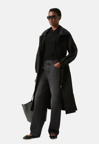 Czarny długi płaszcz na czarnym polo, zestawiony z szarymi szerokimi dżinsami i czarnymi płaskimi butami. Akcesoria to okrągłe czarne okulary przeciwsłoneczne i szara torba.