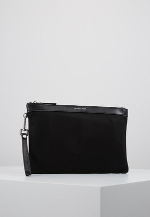 Pochette bracelet noire Michael Kors avec fermeture éclair et sangle en cuir, posée sur une surface blanche sur fond gris.