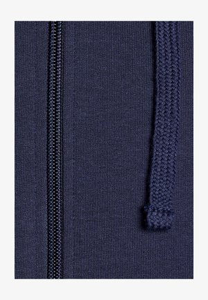 Nahaufnahme von marineblauem Stoff mit einem Reißverschluss auf der linken Seite und einem geflochtenen Kordelzug auf der rechten Seite.