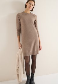 Gebreide sweaterjurk in lichtbruin, met geribbelde halslijn en manchetten. Model houdt een crèmewitte, pluizige jas vast, gecombineerd met zwarte laarzen.