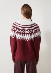 Bordeaux gebreide trui met een witte geribbelde kraag, met een geometrisch ontwerp in wit en lichtroze langs de schouders en de rug.