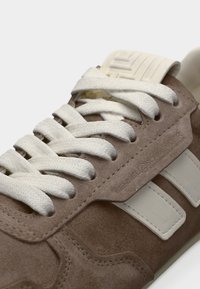 Καφέ σουέντ αθλητικό παπούτσι με λευκά κορδόνια και πλευρικές ρίγες. Διαθέτει υφή στο επάνω μέρος και ενισχυμένη μύτη, με το λογότυπο στο πλάι.