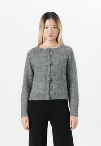 Young woman standing, wearing a buttoned grey textured knit cardigan and loose black pants against a plain white background.