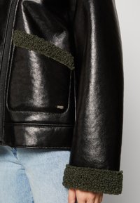Veste en cuir noir avec fermeture éclair, bordure en polaire vert foncé sur les poignets et les poches, avec une texture lisse et brillante.