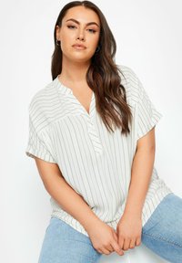 Blusa leggera a maniche corte bianca con righe verticali verdi, un mezzo bottoncino e vestibilità rilassata. Abbinata a jeans blu.