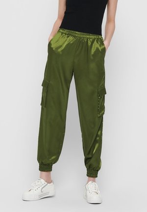 Person iført olivengrønne satin cargo bukser med elastiske manchetter og lommer, kombineret med hvide sneakers og en sort ærmeløs top.