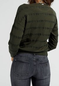 Suéter verde oliva con un patrón texturizado y franjas negras de marca, combinado con jeans gris oscuro con bolsillos traseros y costuras sutiles.