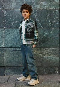 Jongen met krullend haar, gekleed in een groen geruit shirt, een wit T-shirt met opdruk, losse blauwe jeans en beige sneakers, staand tegen een groene marmeren muur.