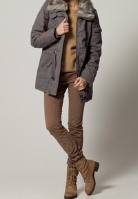Giubbotto imbottito grigio con colletto in pelliccia sintetica, abbinato a pantaloni attillati marrone chiaro e stivaletti in suede con lacci.