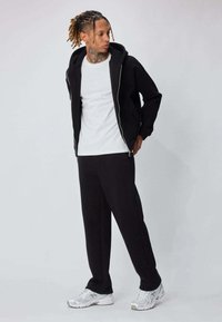 Sweat à capuche noir zippé sur un t-shirt blanc, associé à un pantalon de jogging noir. Porté avec des chaussures de sport blanches. Texture de tissu lisse partout.