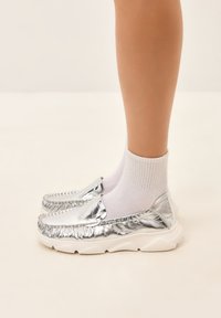 Stříbrné metalické loafers s texturovaným svrškem, bílé robustní podrážky a žebrovanými detaily na okrajích, nošeny s bílými kotníkovými ponožkami.