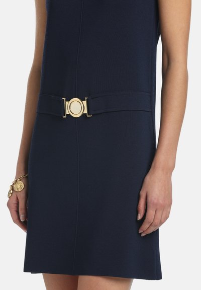 Tmavomodré šaty s priliehavým opaskom v páse so zlatou okrúhlou prackou, nosené so zlatým náramkom s príveskami na ľavom zápästí.