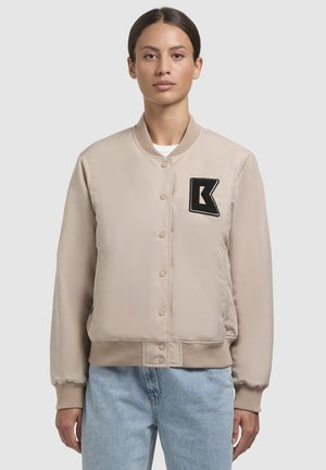 Beige bomber jacket valmistettu sileästä kankaasta, jossa on musta patch tyyliteltyine kirjaimella "K", ribbireunat ja nappikiinnitys.
