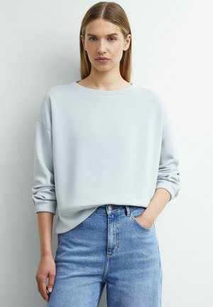 Młoda kobieta z długimi, prostymi włosami, ubrana w jasnoszarą, oversize'ową bluzę i niebieskie jeansy z wysokim stanem, stojąca z jedną ręką w kieszeni.