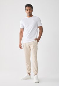 Mann som har på seg en hvit kortermet T-skjorte, beige joggebukser og hvite sneakers, står med den ene hånden i lommen mot en enkel lys bakgrunn.