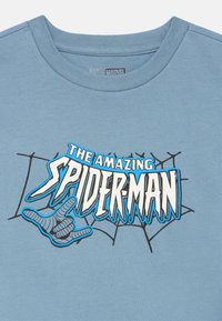 Svetlomodré bavlnené tričko s grafikou "The Amazing Spider-Man" v čiernej a bielej farbe, s motívom ruky a sieťovým dizajnom.
