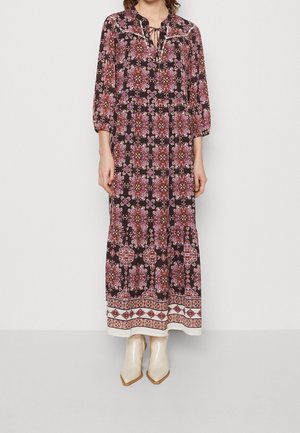 Kobieta ubrana w czarną sukienkę z długim rękawem, sięgającą do kostek, z różowymi kwiatowymi wzorami i białą geometryczną obwódką, w połączeniu z kremowymi, spiczastymi botkami.