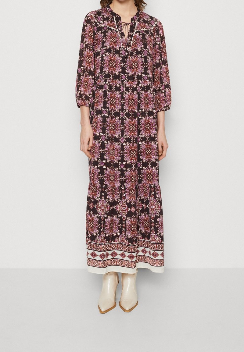 Femme portant une robe noire à manches longues, longue jusqu'aux chevilles, avec des motifs floraux roses et une bordure géométrique blanche, associée à des bottines crème à bouts pointus.