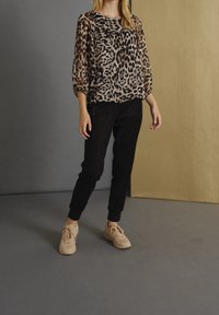 Gepardprint gennemsigtig top med trekvartærmer, parret med sorte tapered bukser og beige sneakers på et gråt gulv mod en gylden baggrund.