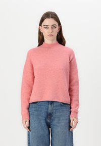 Roza pulover iz mehkega in puhastega materiala z visokim ovratnikom in rebrastimi manšetami. Usklajen z modrimi ohlapnimi jeans hlačami.