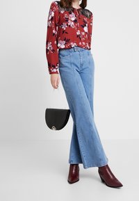 Röd blommig blus med svarta spetsdetaljer, högmidjade blå jeans och bordeauxfärgade klackstövlar. Håller en svart, texturerad handväska.