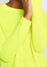 Sweter dzianinowy z długim rękawem w jaskrawym neonowym żółtym kolorze, w luźnym kroju, z szerokim dekoltem i teksturowanym materiałem.