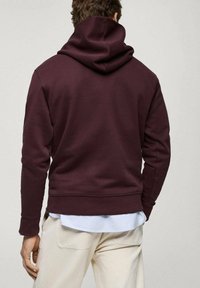 Bordowy hoodie z dużym, miękkim kapturem, ryflowanymi mankietami i dołem, wykonany z materiału mieszanki bawełny. Noszony na jasnoniebieską koszulę z guzikami.