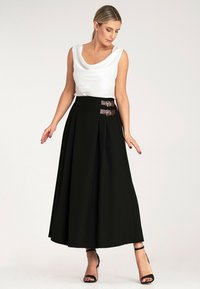 Czarna spódnica plisowana z wysokim stanem i podwójnymi brązowymi klamrami, zestawiona z białą bezrękawnikową bluzką z okrągłym dekoltem.