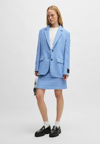 Lichtblauwe gestructureerde blazer met bijpassende minirok, beide met een subtiel patroon. Zwarte knopen en glanzende zwarte instappers maken de look compleet.