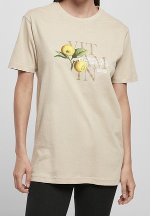 T-shirt beige avec graphique de branche de citron et texte "VITAMIN", porté par une personne avec un pantalon noir, visible du cou aux hanches.