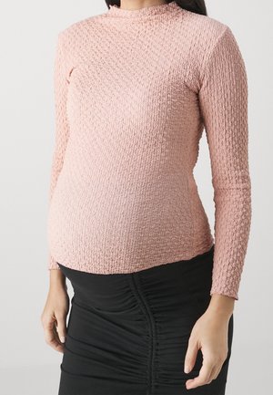 Kvinde iført en tekstureret langærmet lyserød top og en rynket sort nederdel, stående mod en ensfarvet lys baggrund.