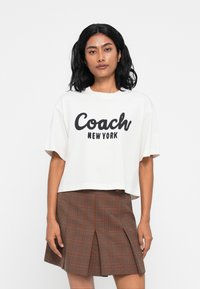 Camiseta blanca recortada con "Coach NEW YORK" en texto negro en negrita. Combinada con una falda de cuadros marrón con pliegues que presenta líneas anaranjadas.