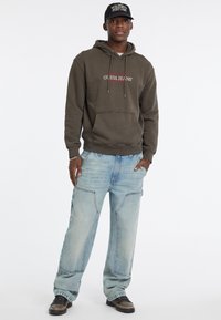 Felpa verde oliva con logo "GUESS JEANS" e tasca frontale. Jeans larghi azzurri con tasche cargo. Scarpe da ginnastica nere con superficie strutturata.