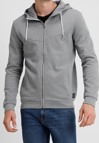 Uomo che indossa una felpa grigia chiara con zip e corde bianche, insieme a jeans blu, in piedi contro uno sfondo semplice.