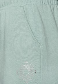 Hellgrüne Strick-Jogginghose mit elastischem Bund, Seiten- und einer silbernen Sonne-Grafik mit radialen Linien auf der unteren Vorderseite.