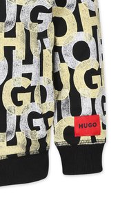 Sweatshirt noir avec un grand design de texte "HUGO" en jaune et blanc, avec une étiquette rouge portant le nom de la marque, des poignets côtelés et un tissu doux.