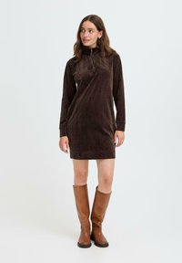 Femme portant une robe en velours côtelé marron à manches longues avec une fermeture éclair sur le devant, associée à des bottes en cuir marron jusqu'aux genoux, debout et regardant sur le côté.