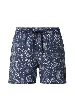 Wysoki umięśniony mężczyzna w granatowych kąpielówkach z wzorem i czarnych klapkach, stojący przodem na jednolitym tle.