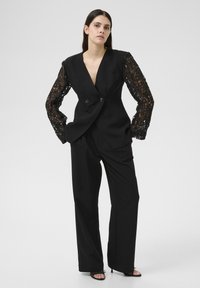 Sort skræddersyet blazer med lange sorte blonde ærmer, dobbelte knapper og matchende vide bukser, præsenteret mod en neutral baggrund.