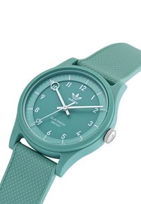 Montre solaire turquoise avec un bracelet en silicone texturé, boîtier rond, chiffres arabes et aiguilles blanches. Elle possède une couronne sur le côté.